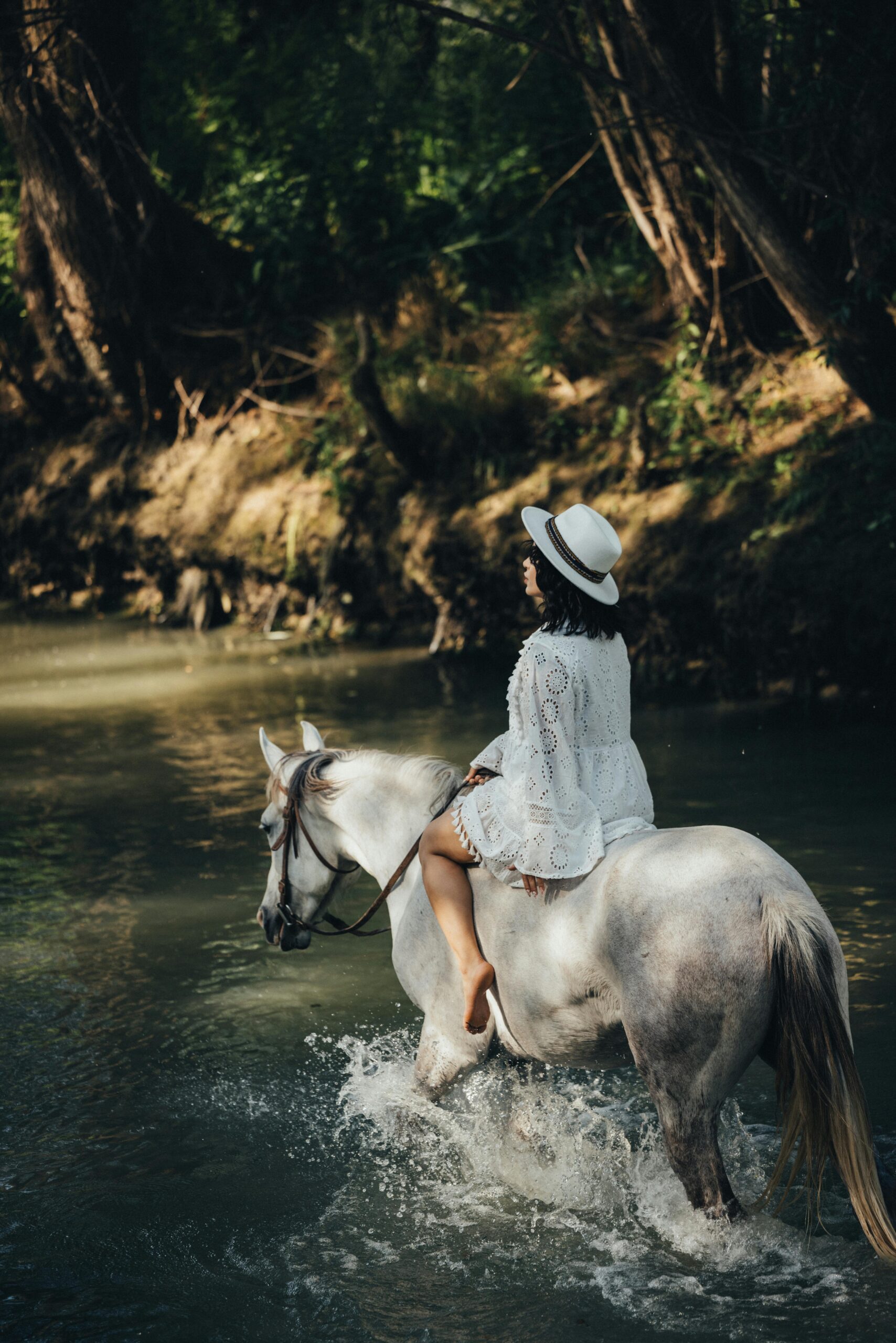 A woman in a white dress rides a horse through a peaceful forest stream.