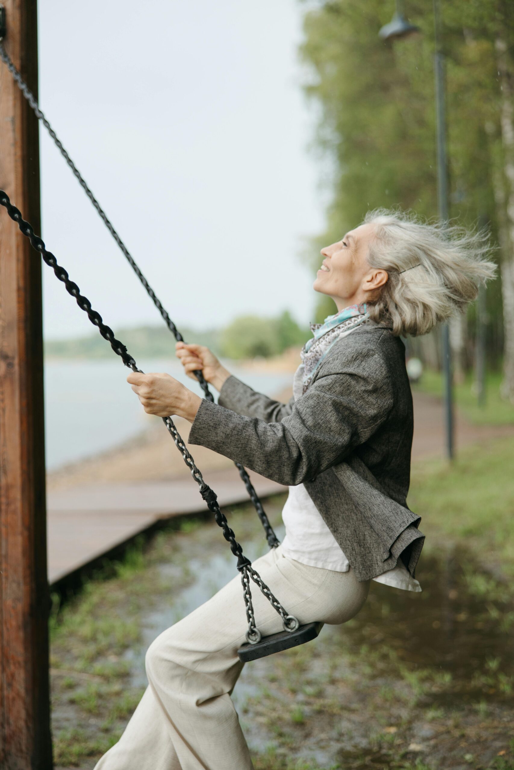 A joyful senior woman swinging in a park, surrounded by nature on a bright day.
